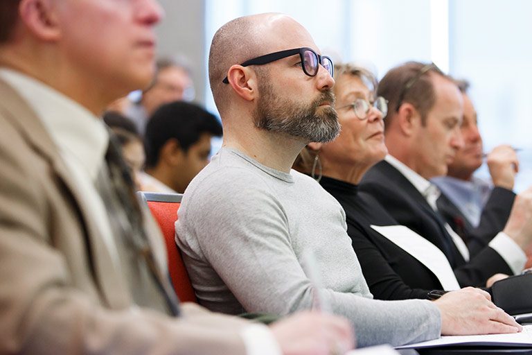 Professionals sit in rows of seats, take notes and look toward the front of the room during an event.