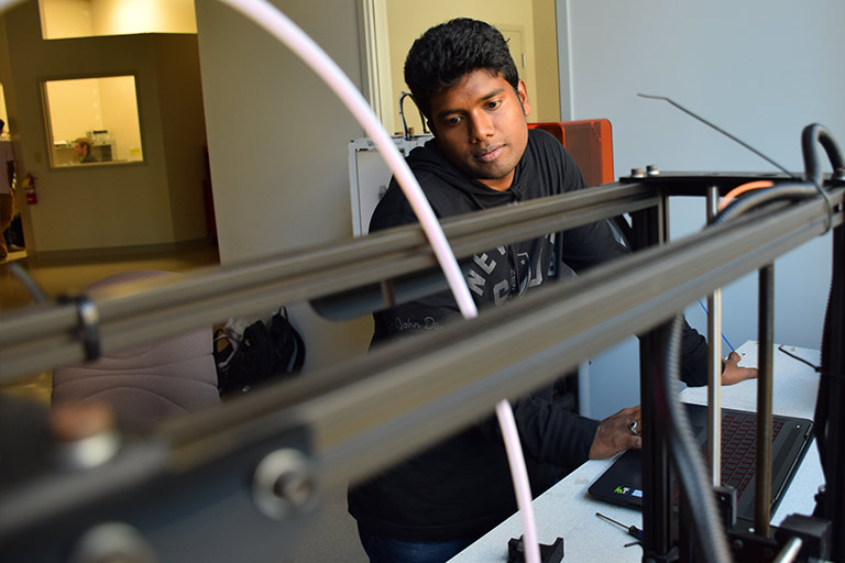 A student stands at a computer desk as he works with a large 3D printer.