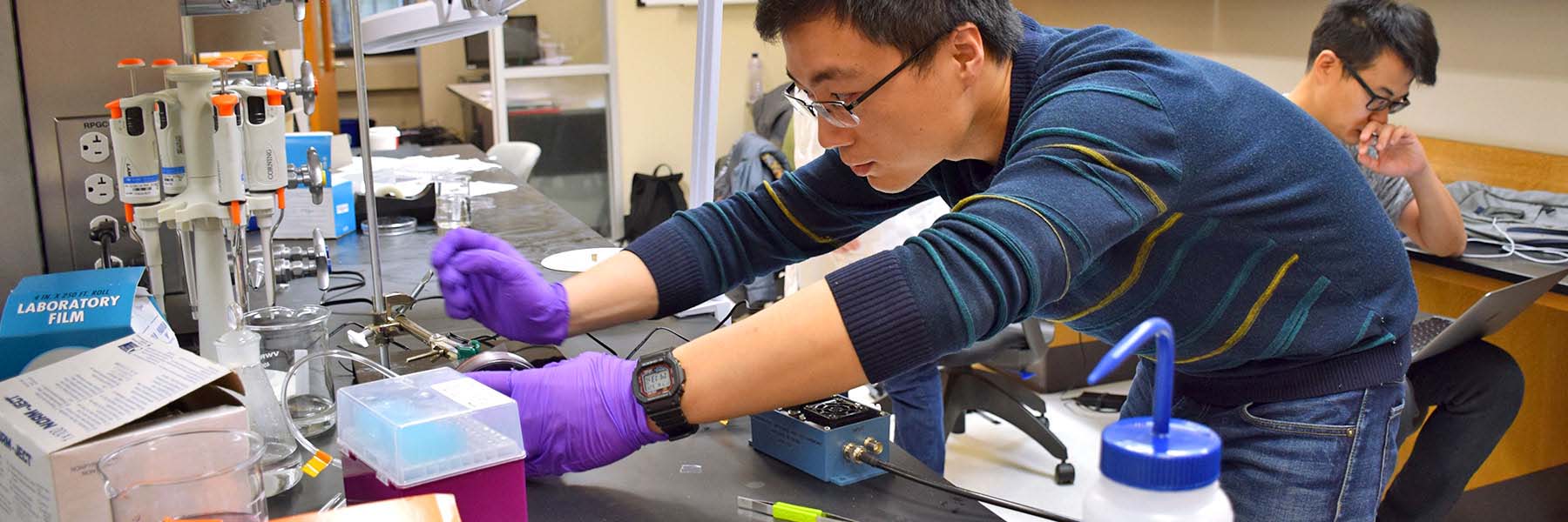 A researcher wears gloves and works with lab equipment while another researcher works on a laptop.