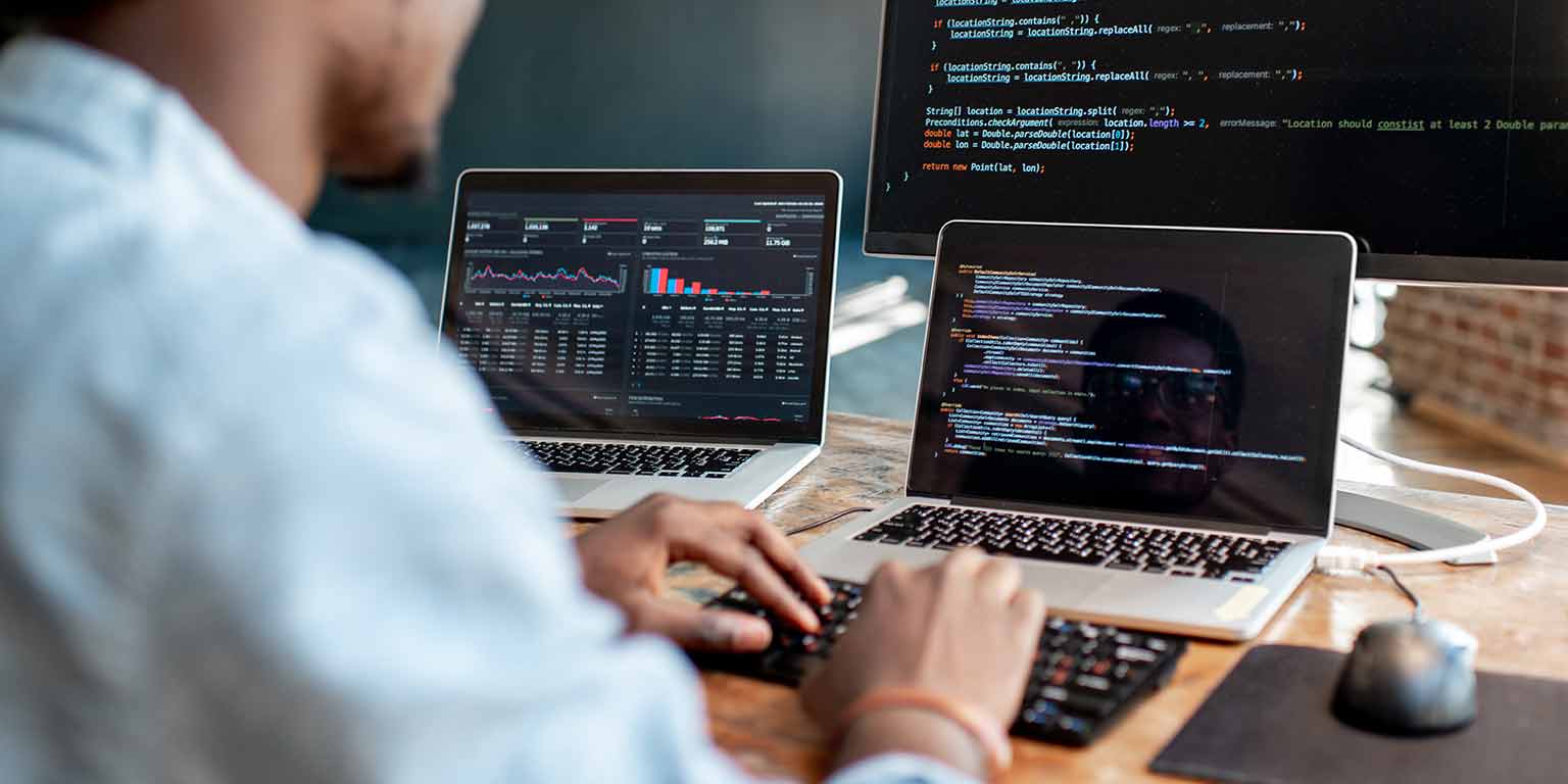 A person writes code on a laptop with another laptop and a desktop monitor sitting on his desk.