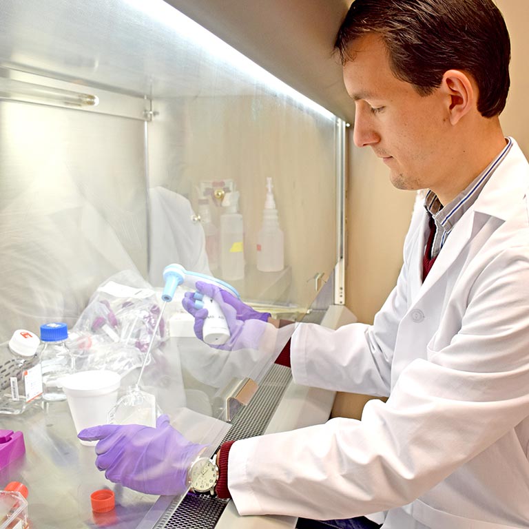 A researcher in a lab coat uses equipment to fill a container with liquid behind a screen.